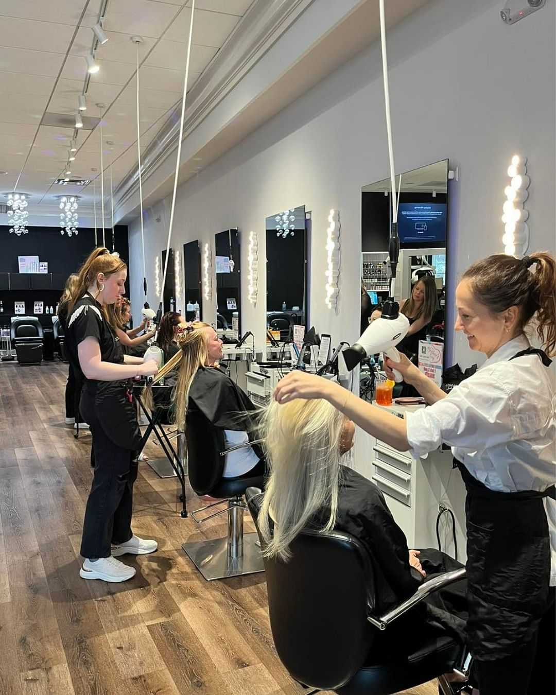 Stylists working on clients' hair in a modern salon with large mirrors and bright lights.