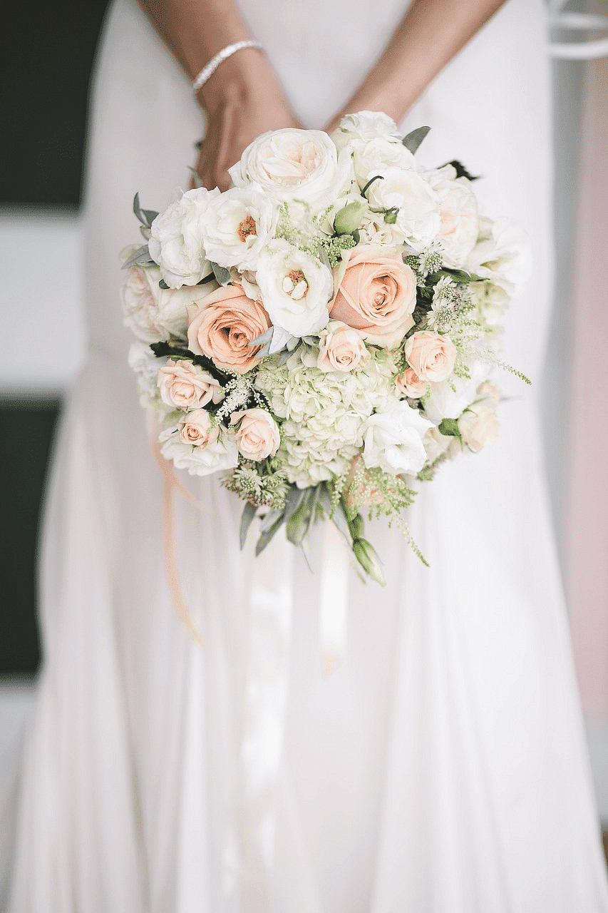 Bride holding a bouquet of white and peach roses against a white wedding dress.