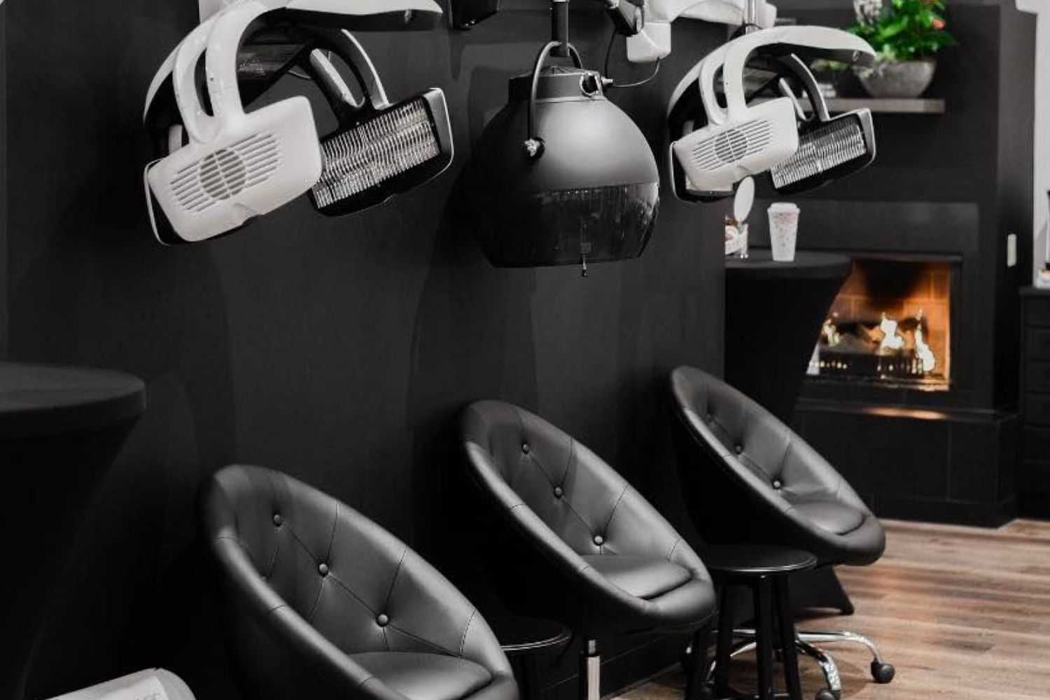 Modern salon with black chairs, overhead hair dryers, and a fireplace in the background.