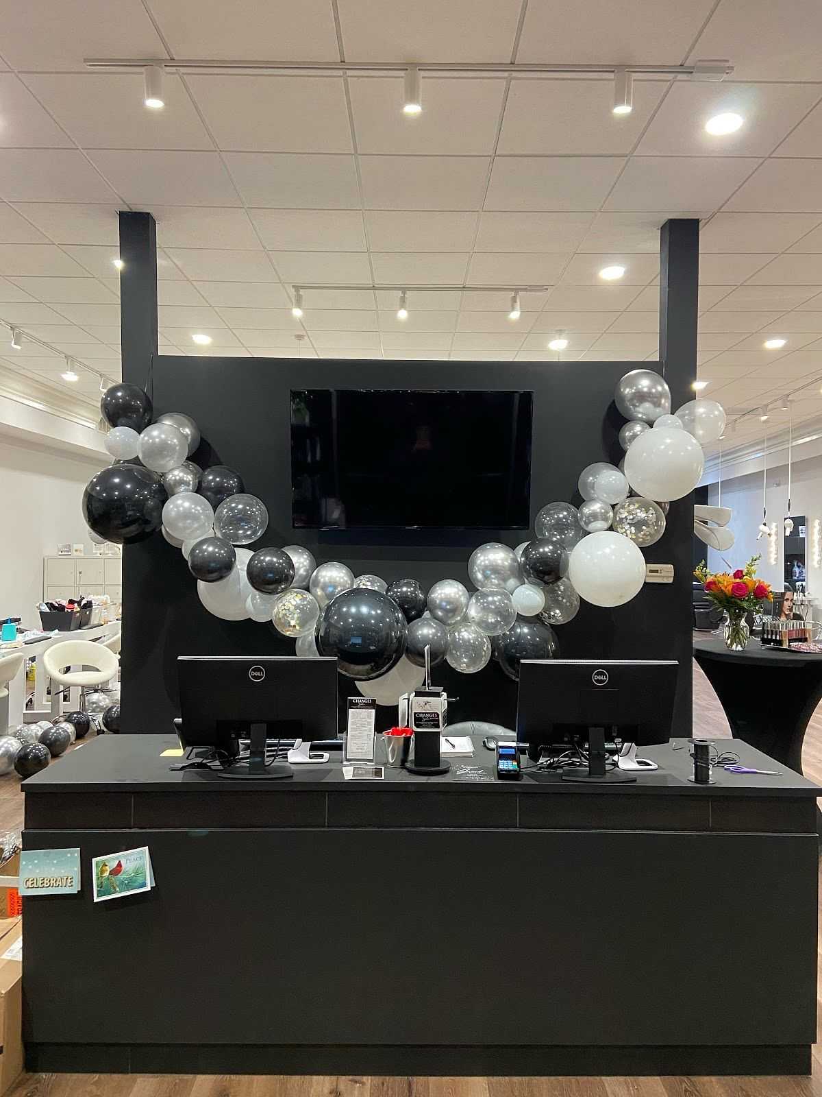 Lobby desk decorated with black, white, and silver balloons, two monitors, and flower bouquet on side table.